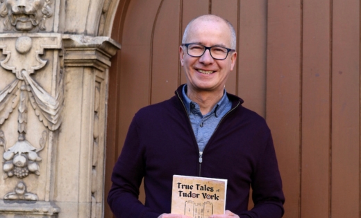 Tony Morgan holding a copy of his book 'True tales of Tudor York', standing outside an historic wooden door