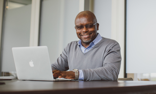 A stock photo of a smiling man sitting at a laptop. July 2020.