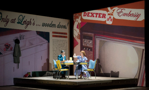 A still from the Opera North production of Trouble in Tahiti, with two actors talking over breakfast in front of two large, retro adverts.
