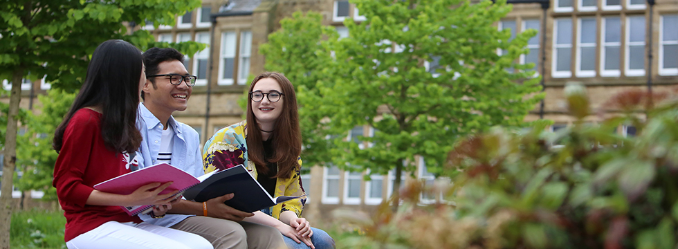 Three smiling students sat outside on a bench in front of a University of Leeds building
