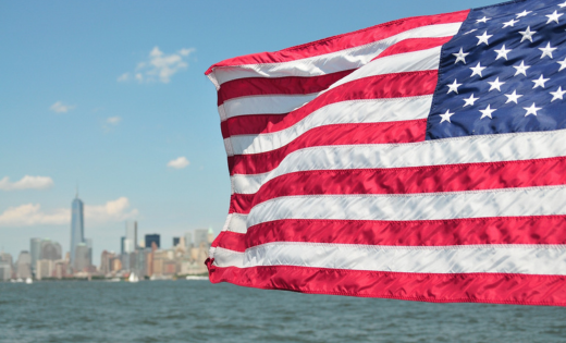 The United States flag flying against the backdrop of a city next to a lake, and a blue sky.